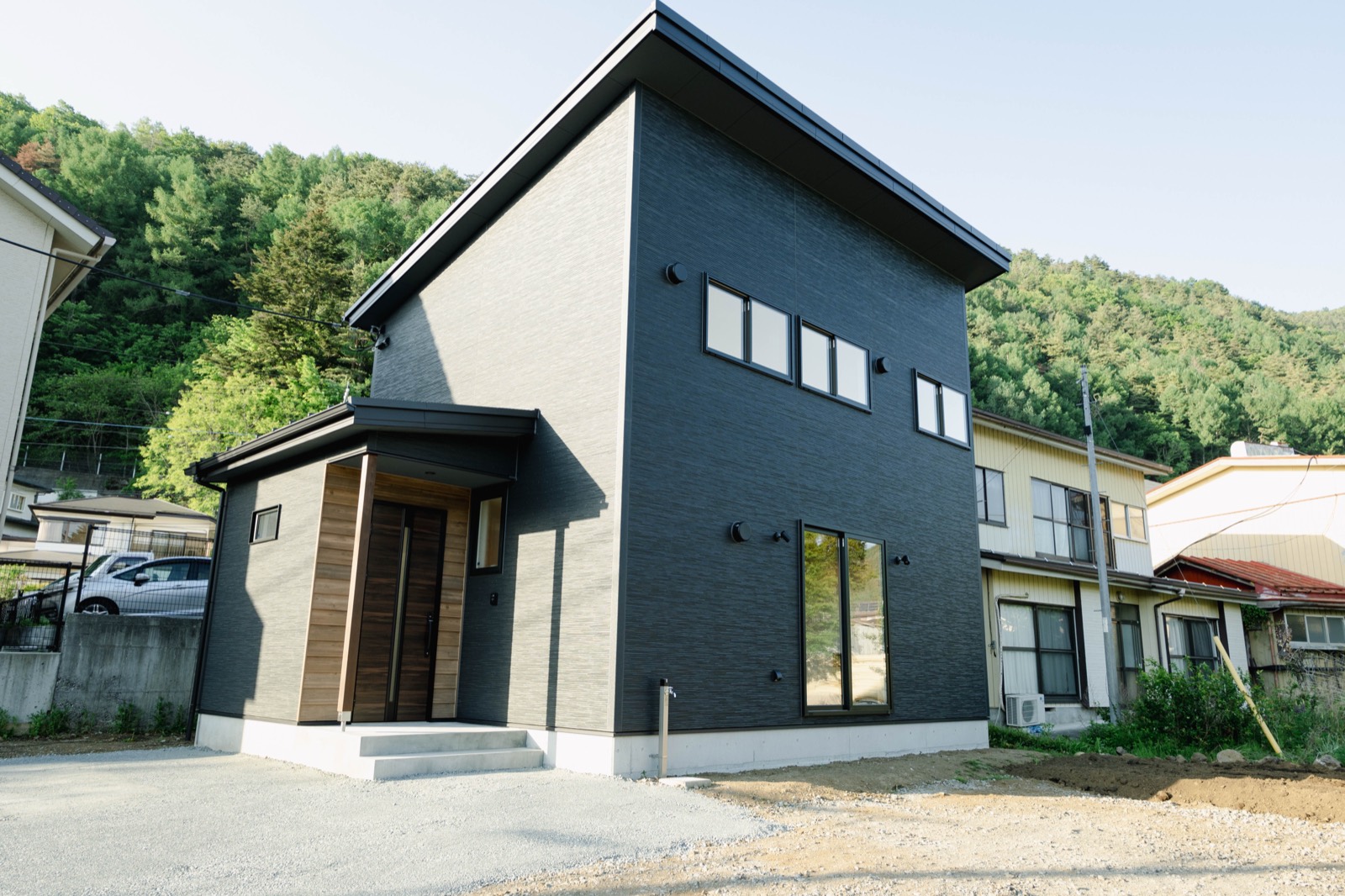 片流れ屋根と黒い外壁でまとめた新築住宅の外観。背景に山の緑が広がる